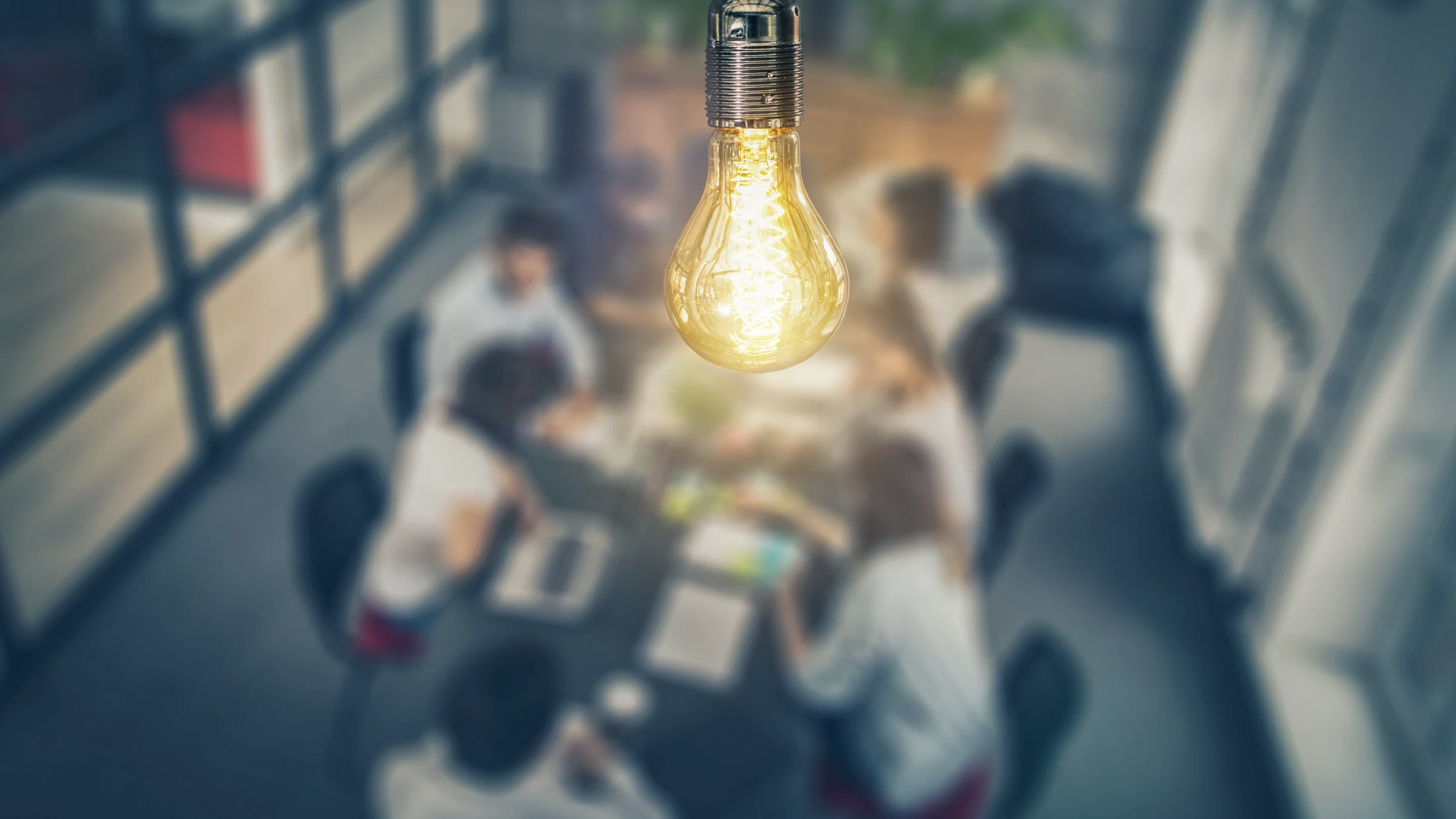 Top-down view of a dynamic business meeting with six participants. The surroundings are intentionally blurred, drawing attention to a vibrant yellow light bulb emitting a brilliant beam of light.